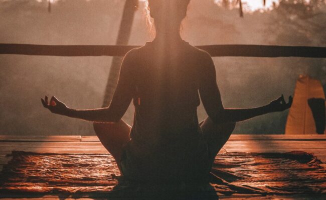 Frau sitzt vor einer Palme im Sonnenuntergang und meditiert
