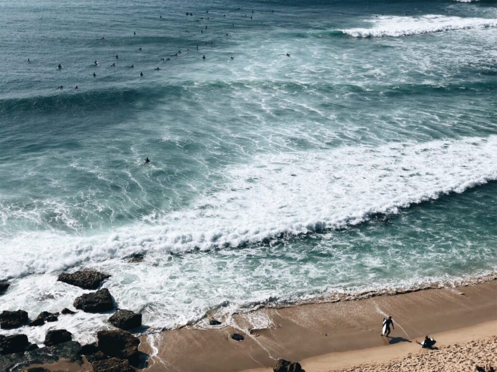 Der Strand von Ericeira in Portugal von oben fotografiert. Es sind viele Surfer im Meer zu sehen.